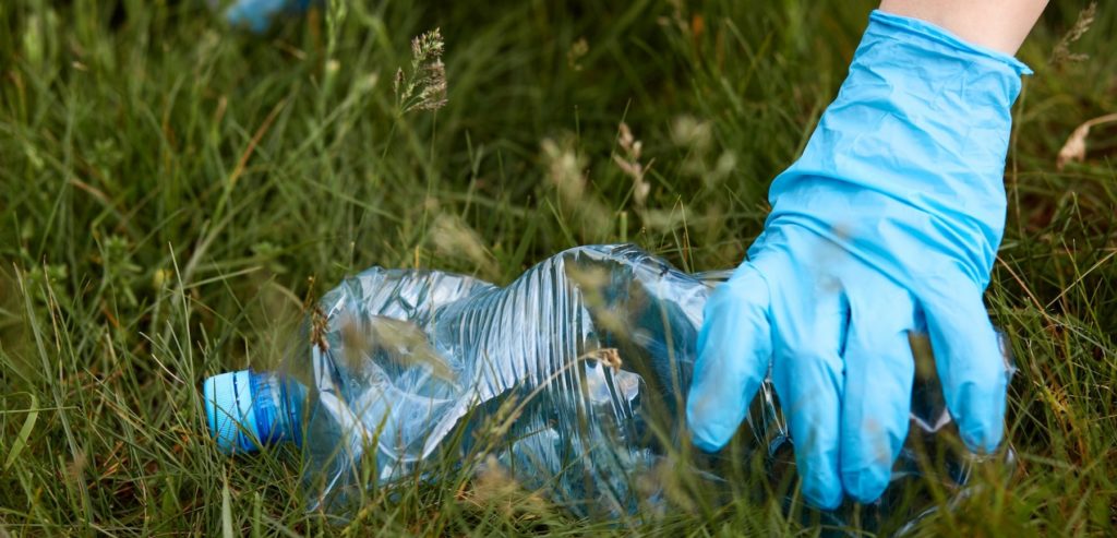 Une main avec un gant d'une personne qui ramasse une bouteille plastique vide dans l'herbe. Cela représente une action du World Clean Up Day