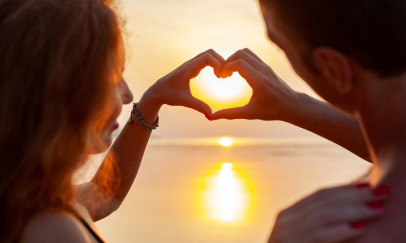 Un jeune couple au bord de l'eau au coucher de soleil qui forme un coeur avec leurs mains