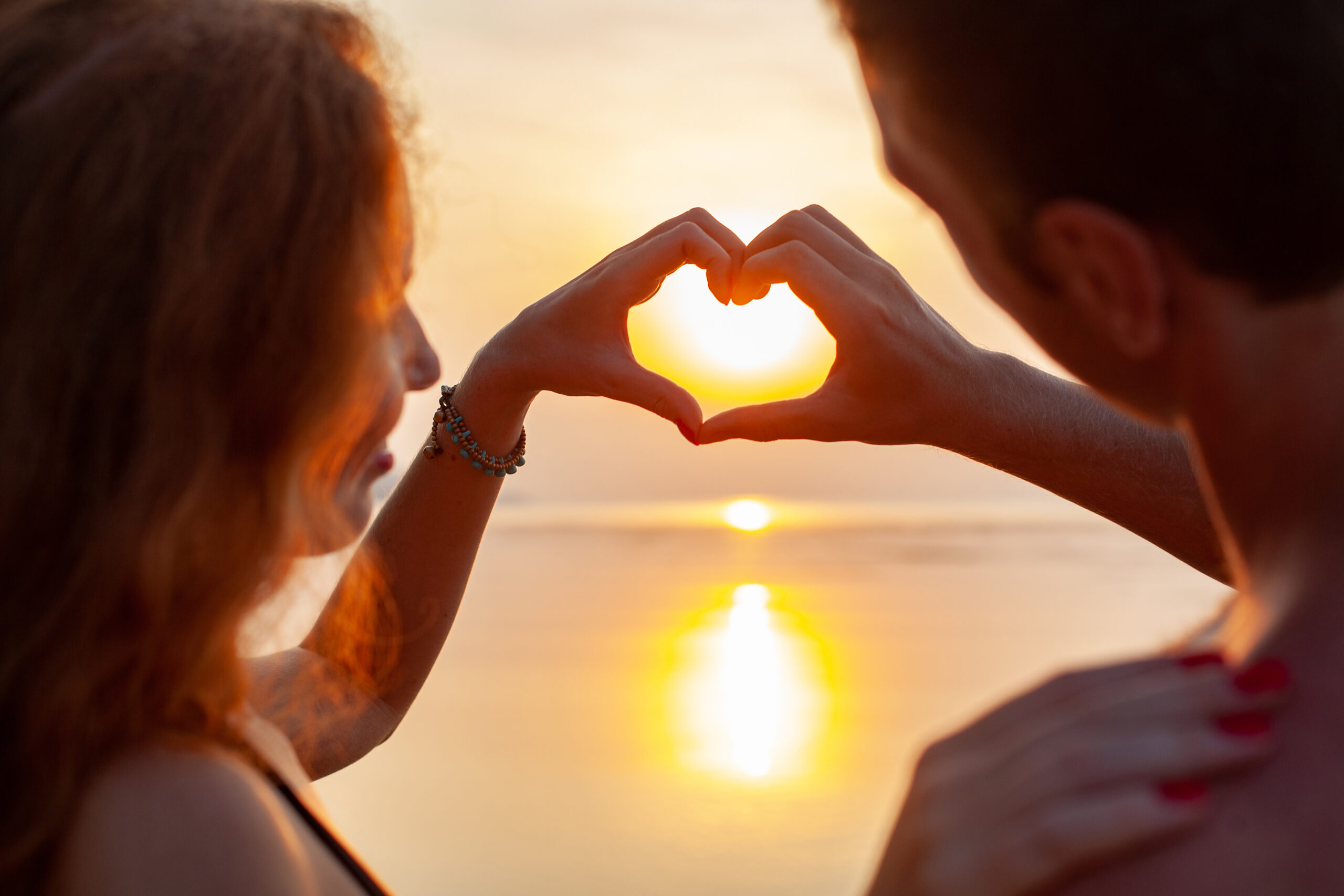 Un jeune couple au bord de l'eau au coucher de soleil qui forme un coeur avec leurs mains